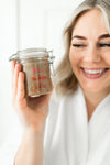 smiling woman holding up a container of body and face sugar scrub for smoothing skin